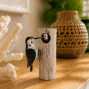 An image of a black and white wooden hand carved and hand painted woodpecker on wood with a baby peering through
